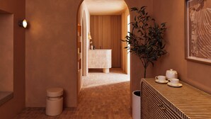 Un pasillo que conduce a un baño con lavabo, espejo y perchero. Hay una planta en maceta y un cuadro en la pared.