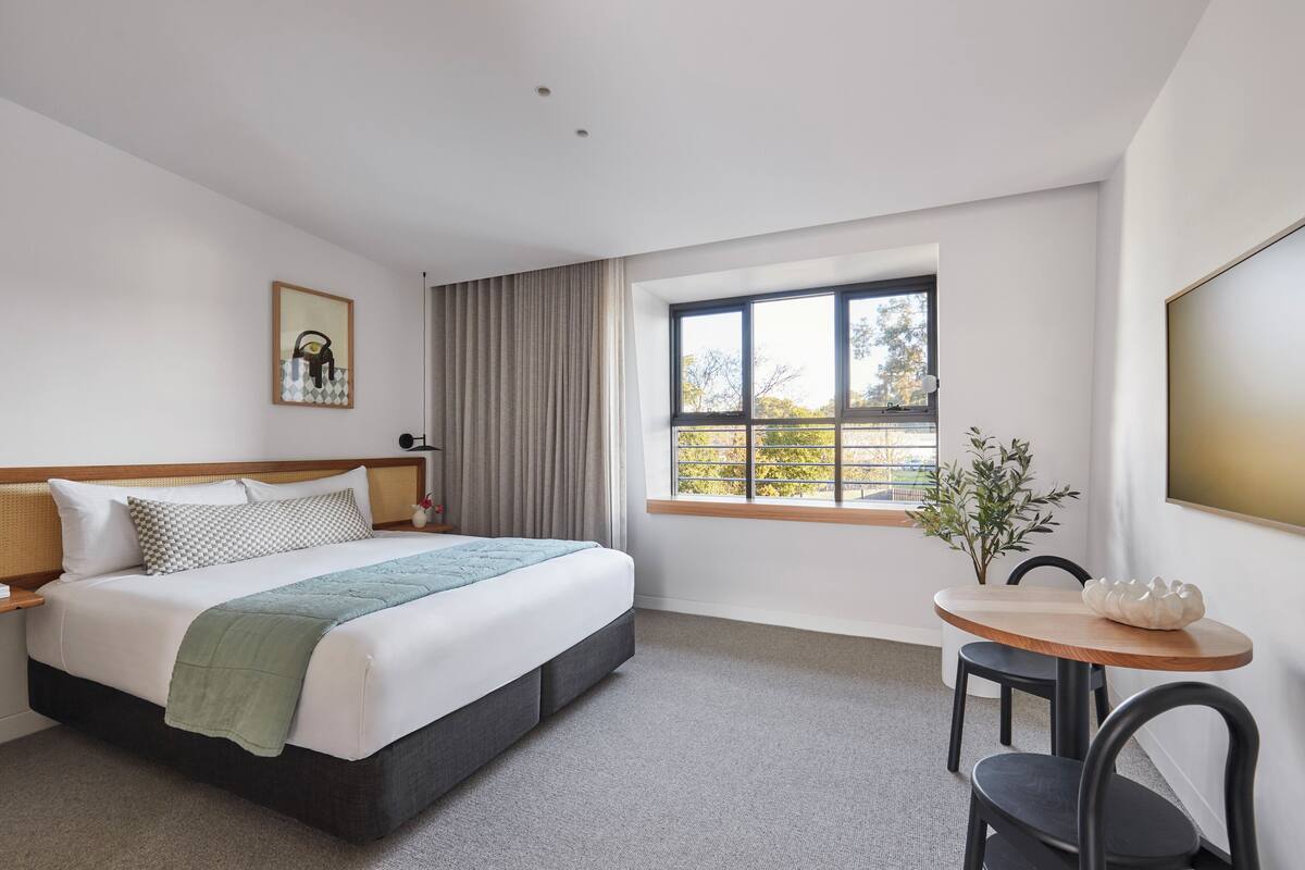 A hotel room with a bed, a window with curtains, a small table with chairs, and a wall-mounted TV.