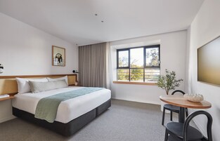 Habitación de hotel con cama, ventana con cortinas, una mesita con sillas y un televisor montado en la pared.