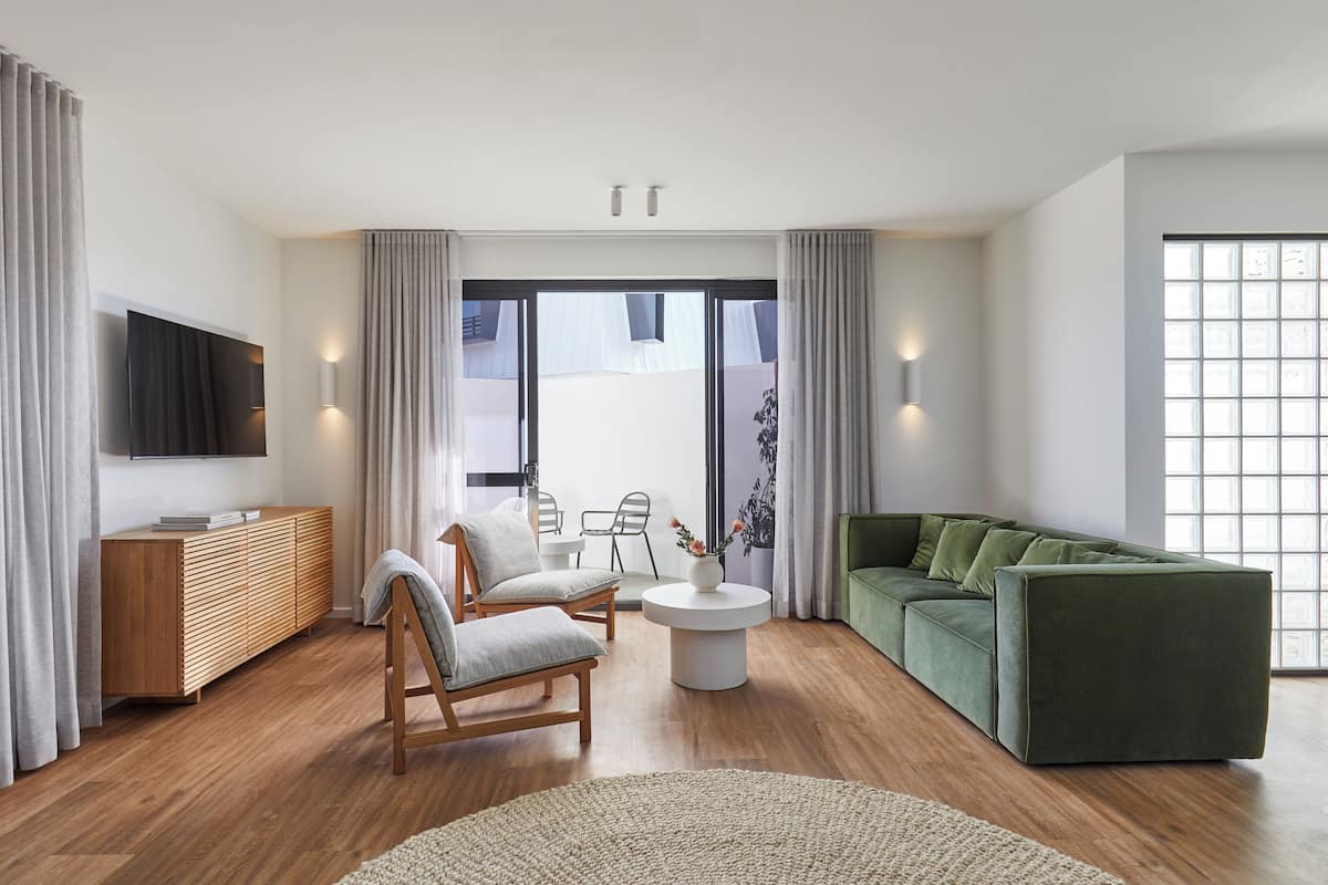 A modern living room with a wooden floor, a green sofa, a wooden cabinet, a TV mounted on the wall, and a large window with curtains.