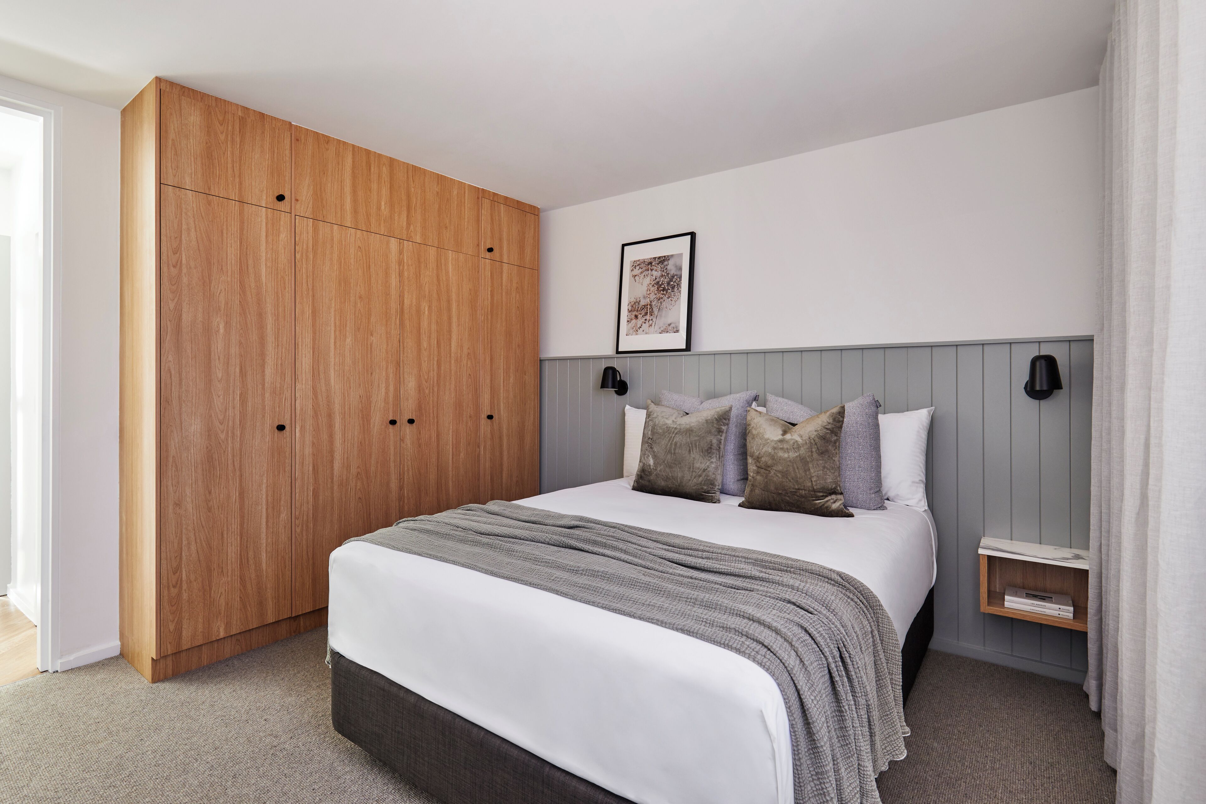 Een slaapkamer met een grote houten garderobe, een bed met een grijze deken en een nachtkastje met een lamp.