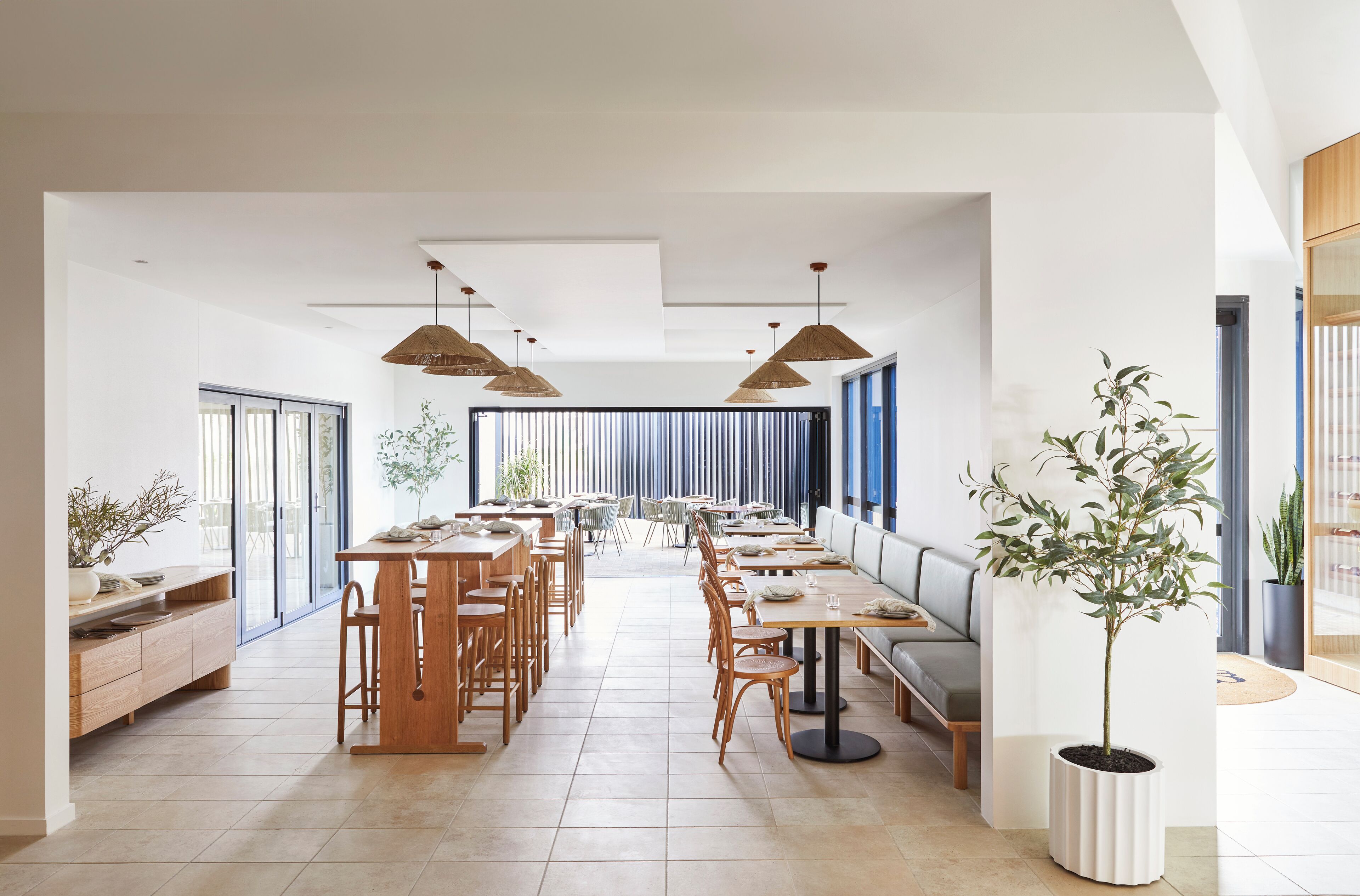 Un vaste espace dédié à la salle à manger, avec de longues tables et des chaises en bois, de grandes fenêtres et des luminaires suspendus.