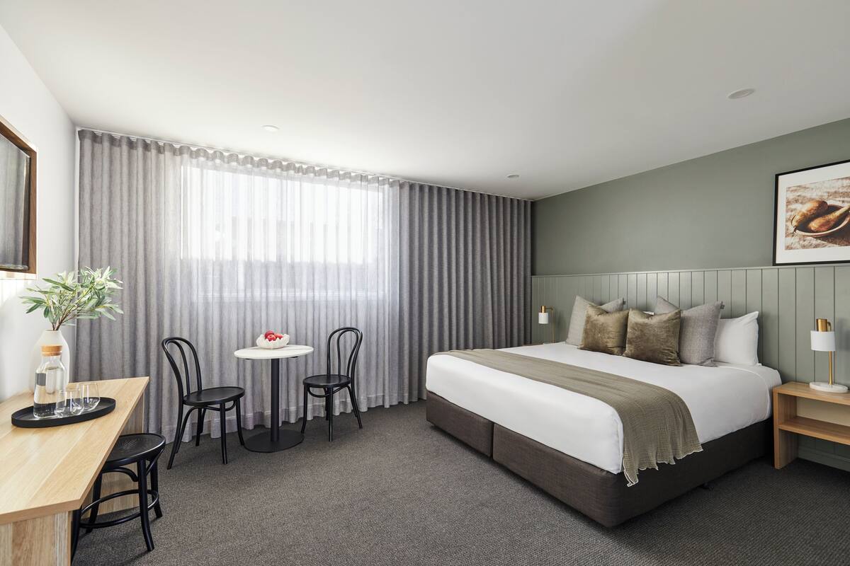 A hotel room with a large bed, a desk with chairs, a small table, and a window with curtains.
