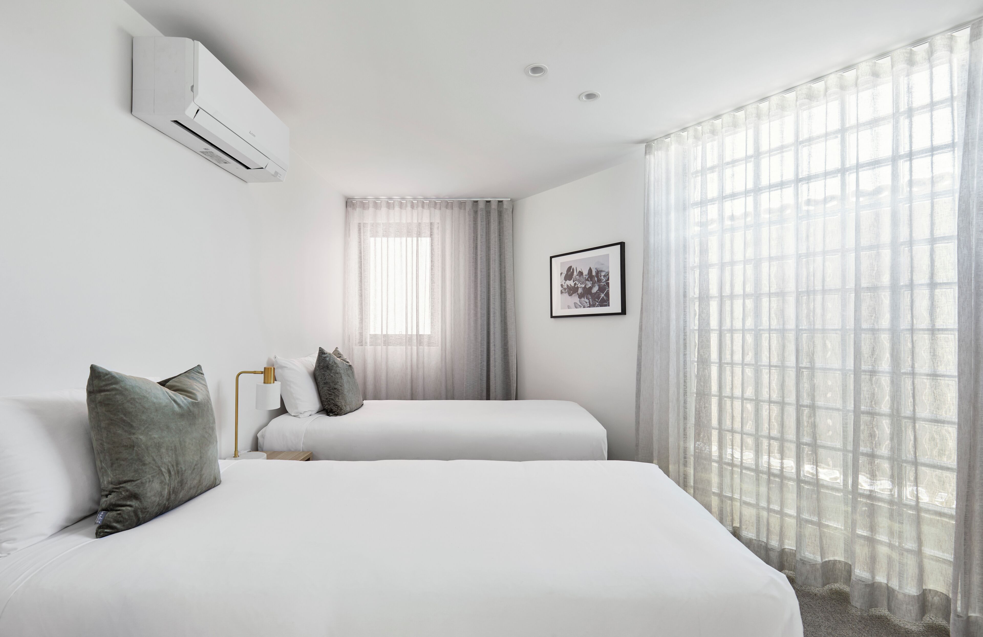 Een hotelkamer met twee bedden, een raam met dunne gordijnen, een wandairconditioner en een ingelijst kunstwerk aan de muur.