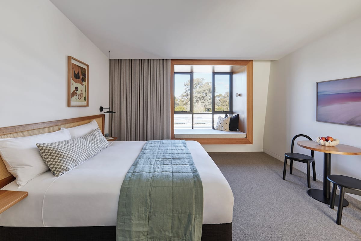 A hotel room with a large bed, a window with a view, a small table, and a chair.