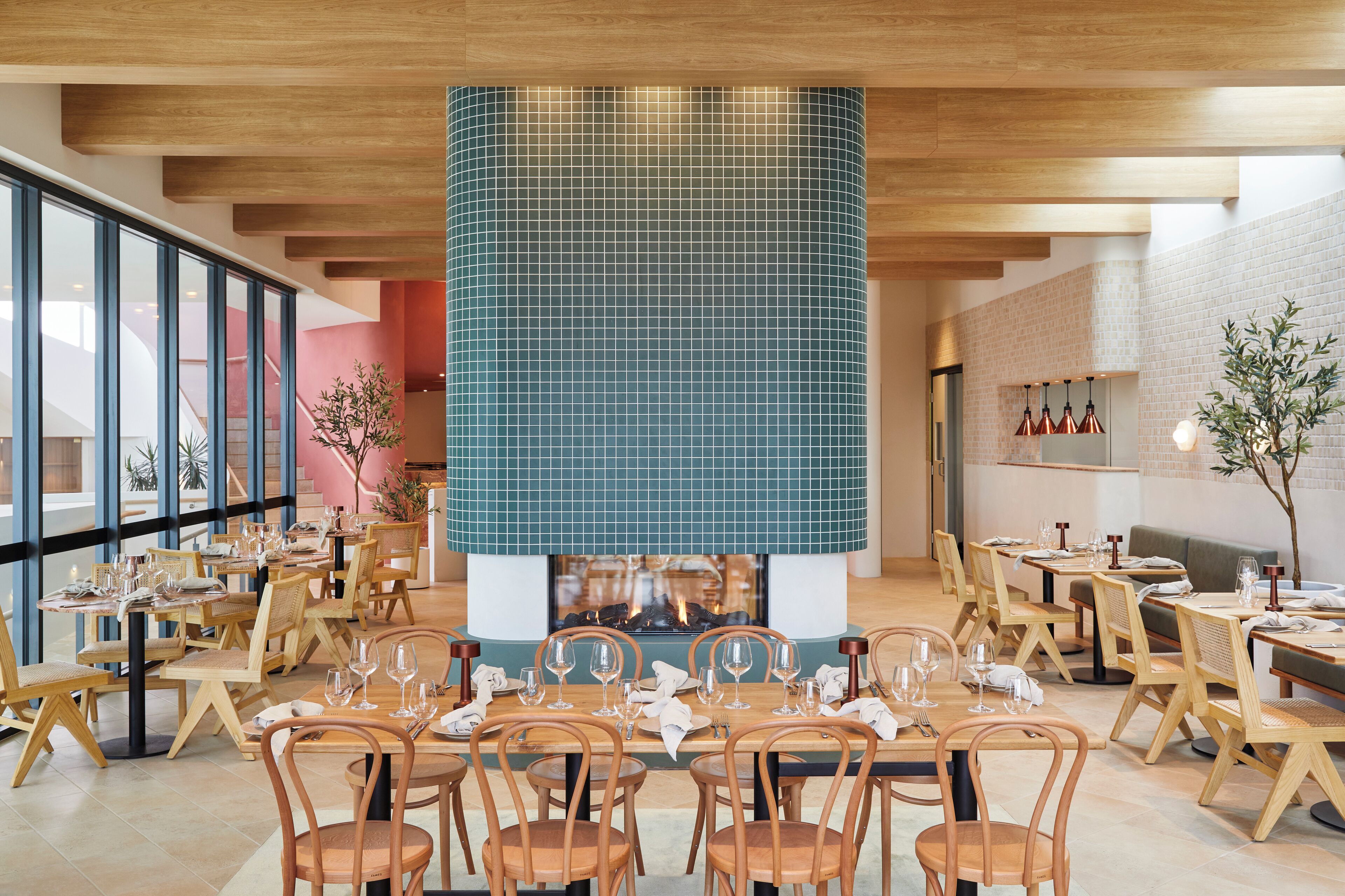 A restaurant with wooden tables and chairs, a central fire feature, and large windows.