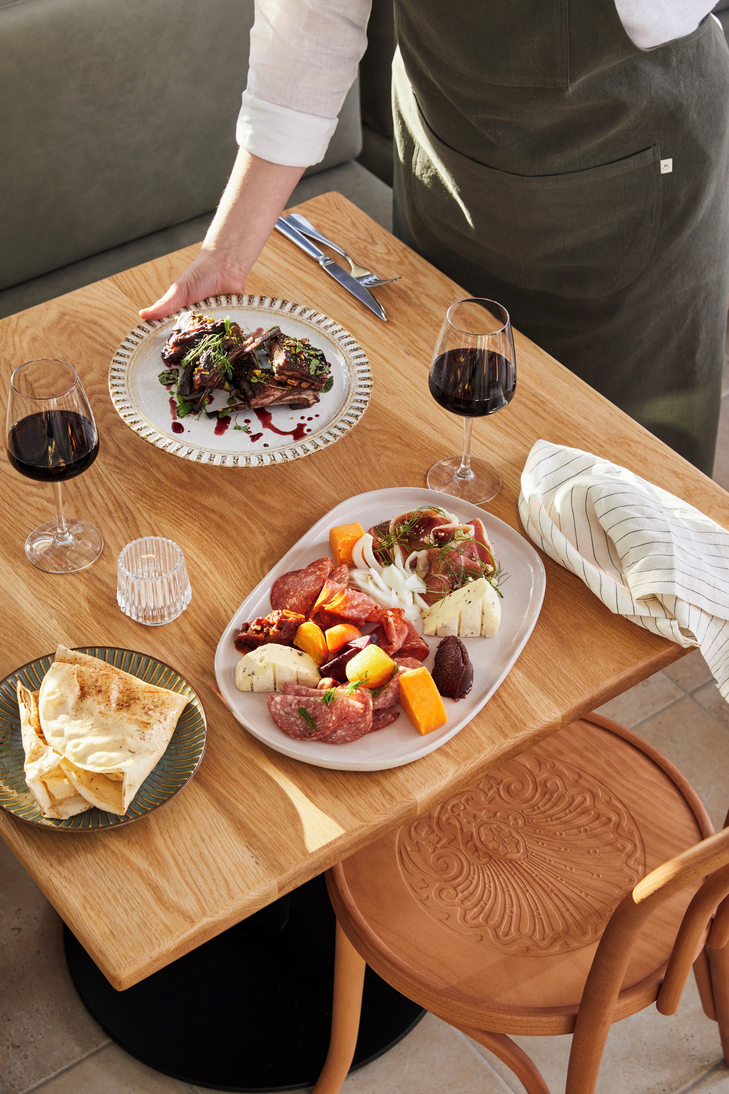 Una mesa de madera con platos de comida, copas de vino y una persona con delantal.