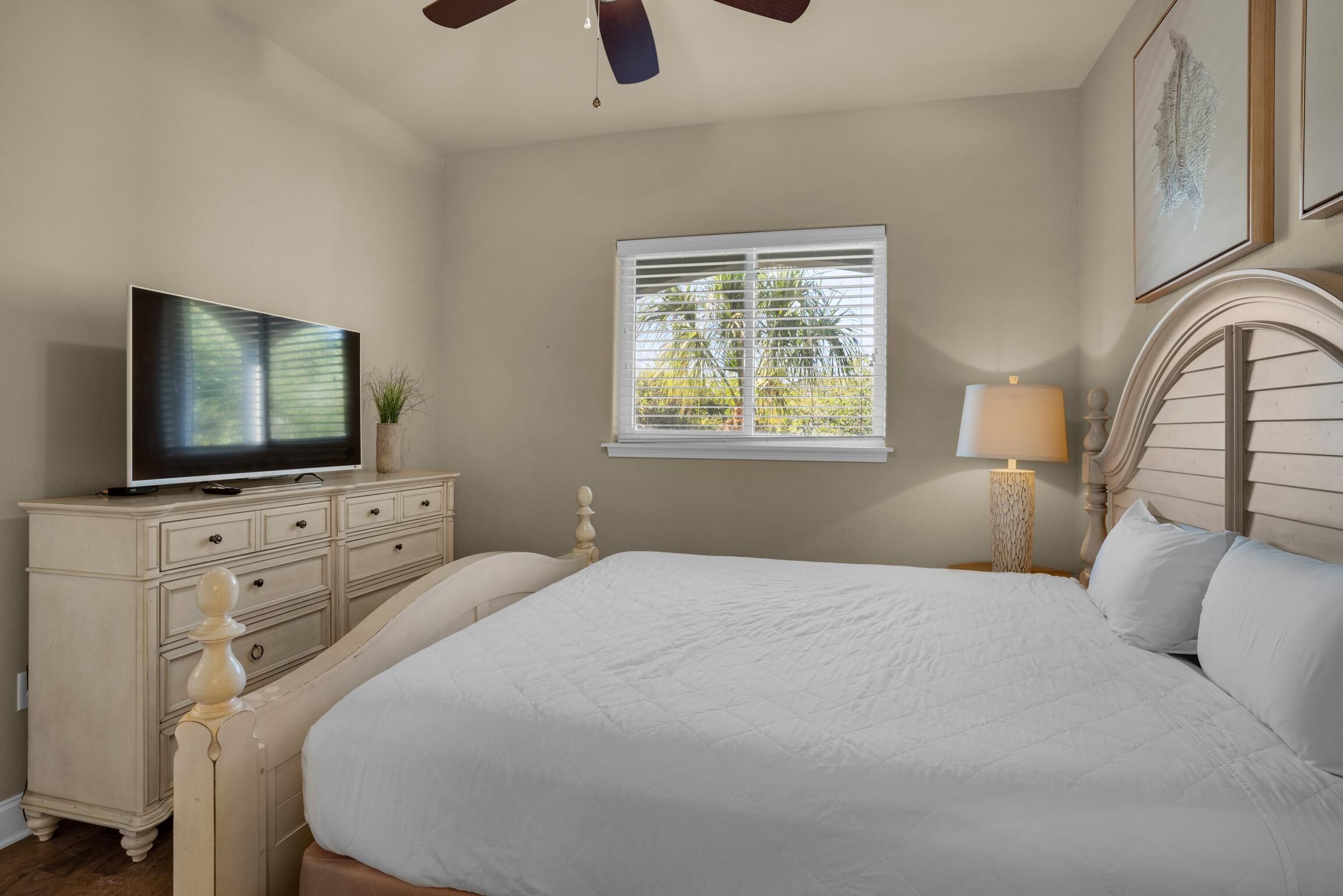 A bedroom with a bed, a TV on a dresser, a window with blinds, and a lamp.