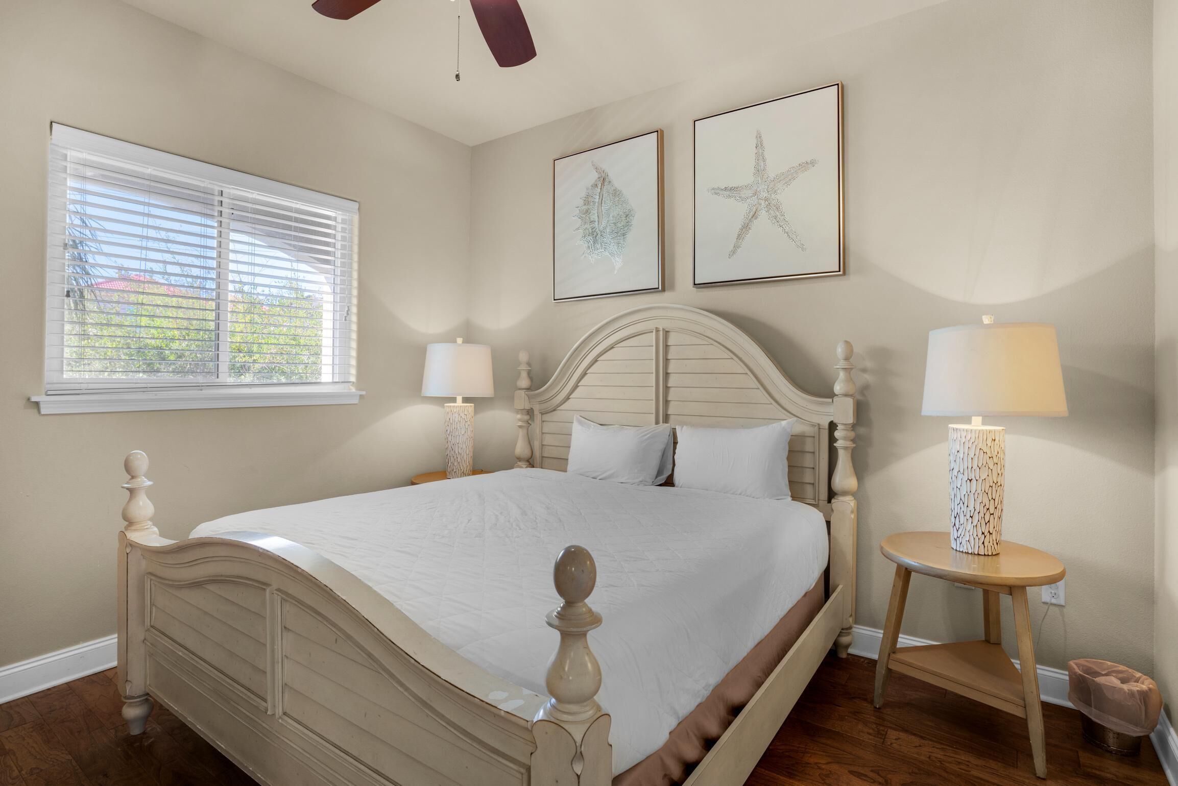 A bedroom with a large bed, two bedside lamps, a window with blinds, and two framed pictures on the wall.