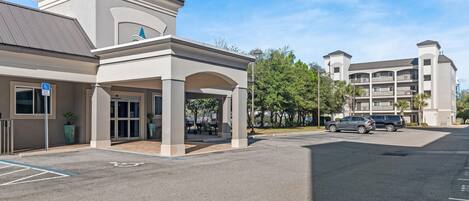 A hotel entrance with a covered walkway, a parking lot, and a multi-story building in the background.