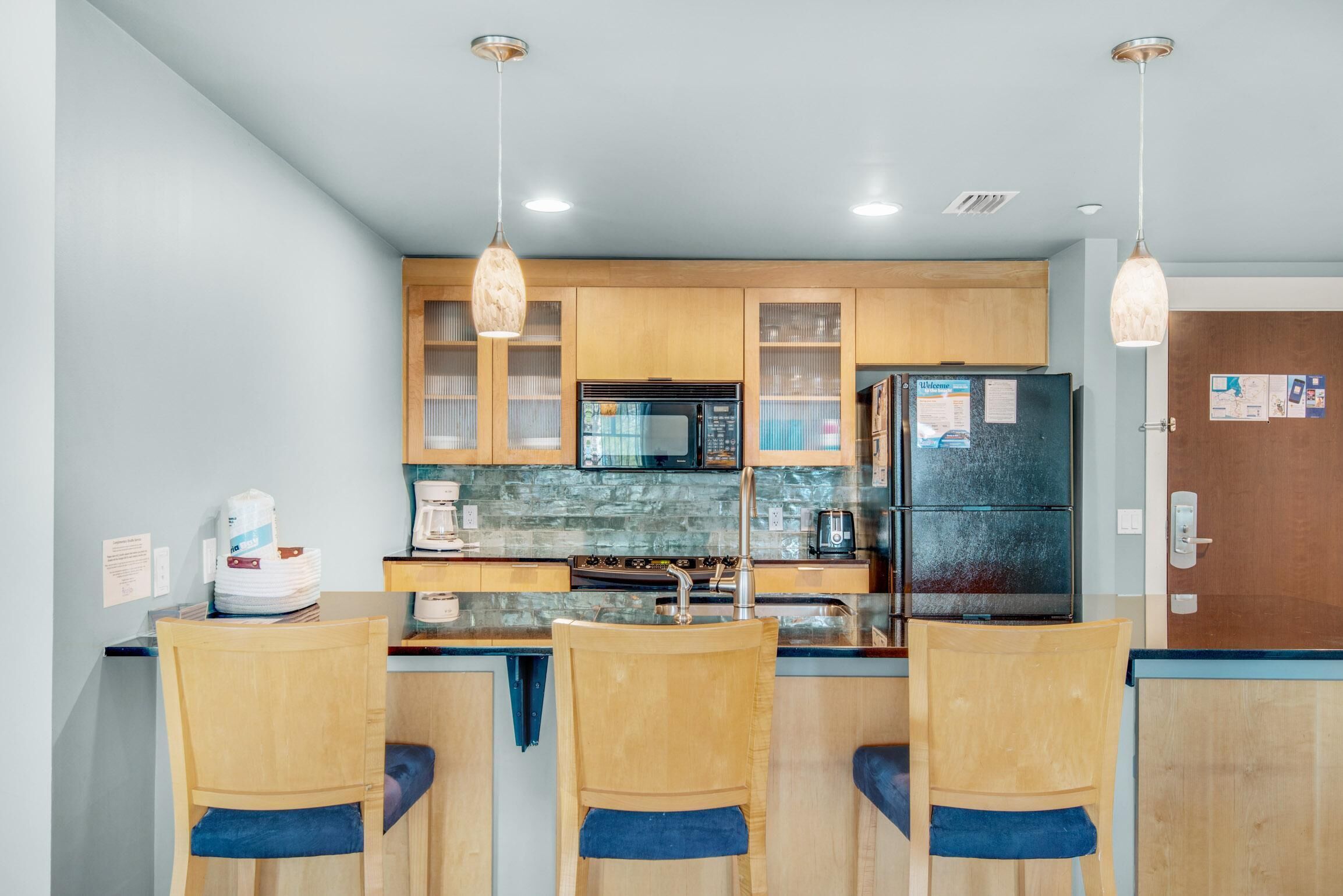 A modern kitchen with wooden cabinets, a black refrigerator, a microwave, and a sink. There are three wooden chairs with blue cushions.