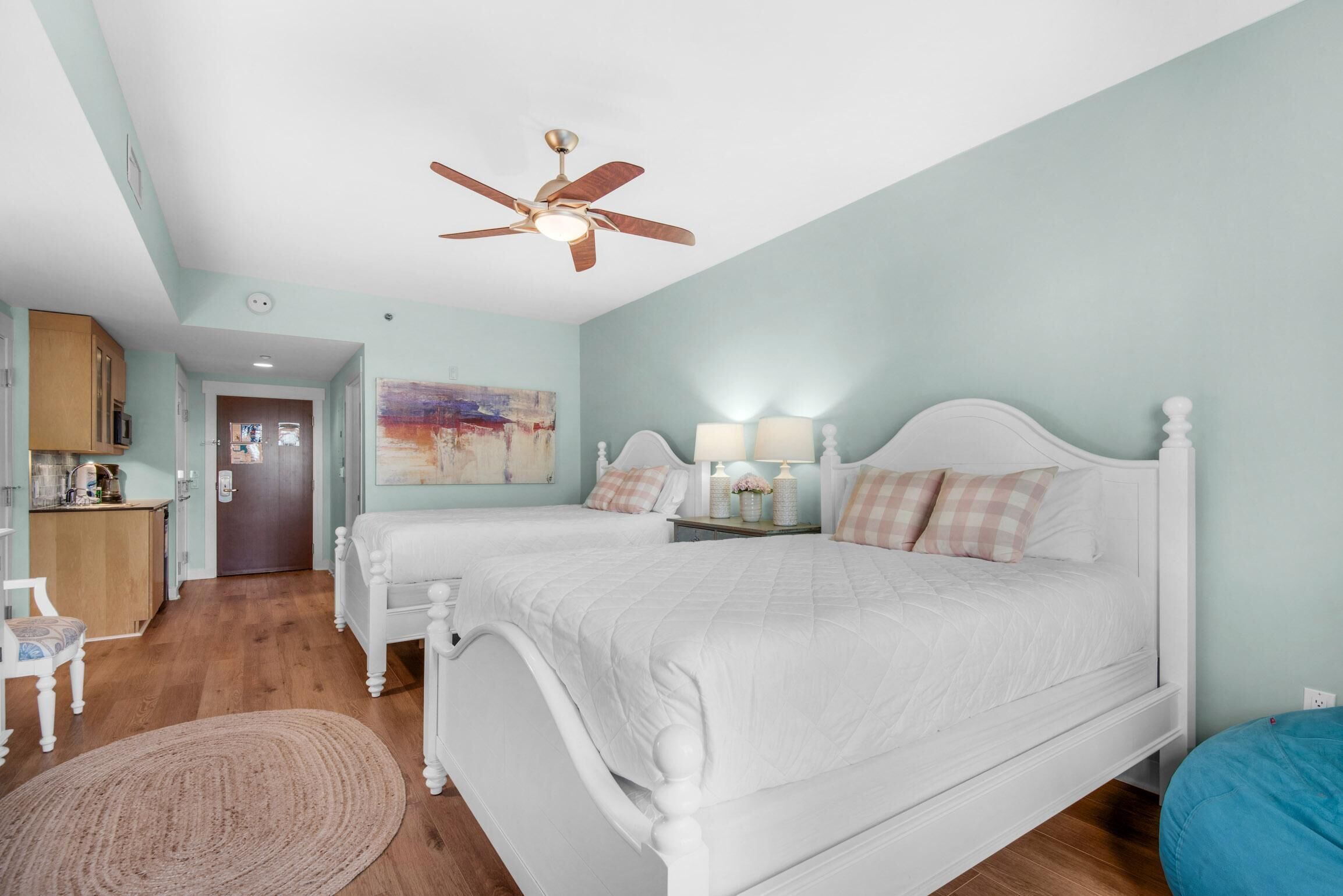 A bedroom with two white beds, a ceiling fan, a painting on the wall, and a wooden floor.