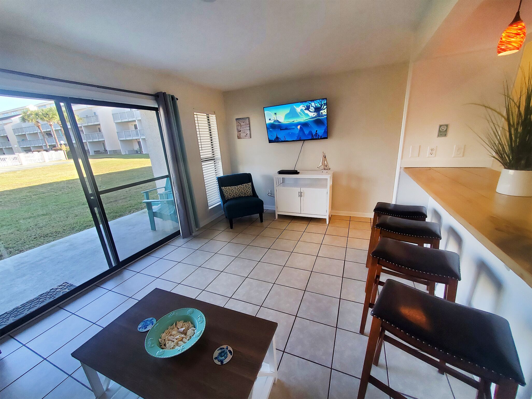A modern living room with a TV, a dining area with chairs, a table with a bowl, and a balcony with a view of buildings and a lawn.