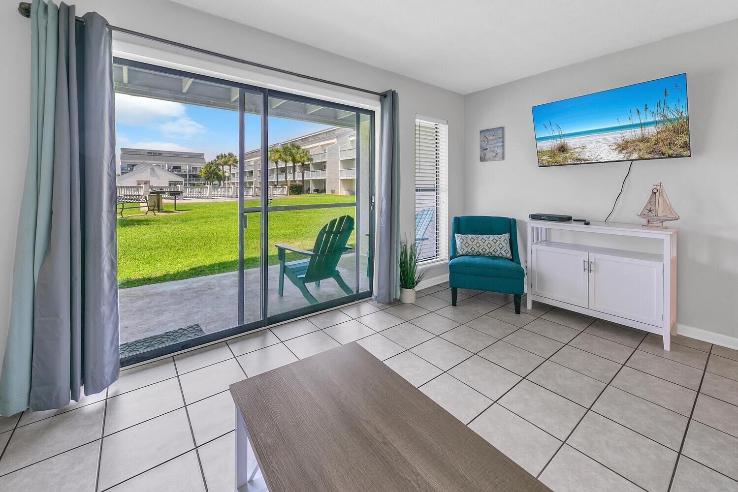 A modern living room with a sliding glass door leading to a balcony, a teal armchair, a white console table, and a painting of a beach scene.