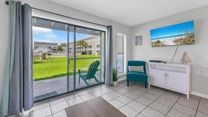 A modern living room with a sliding glass door leading to a balcony, a teal armchair, a white console table, and a painting of a beach scene. - Endless Summer B4 (Panama City Beach)