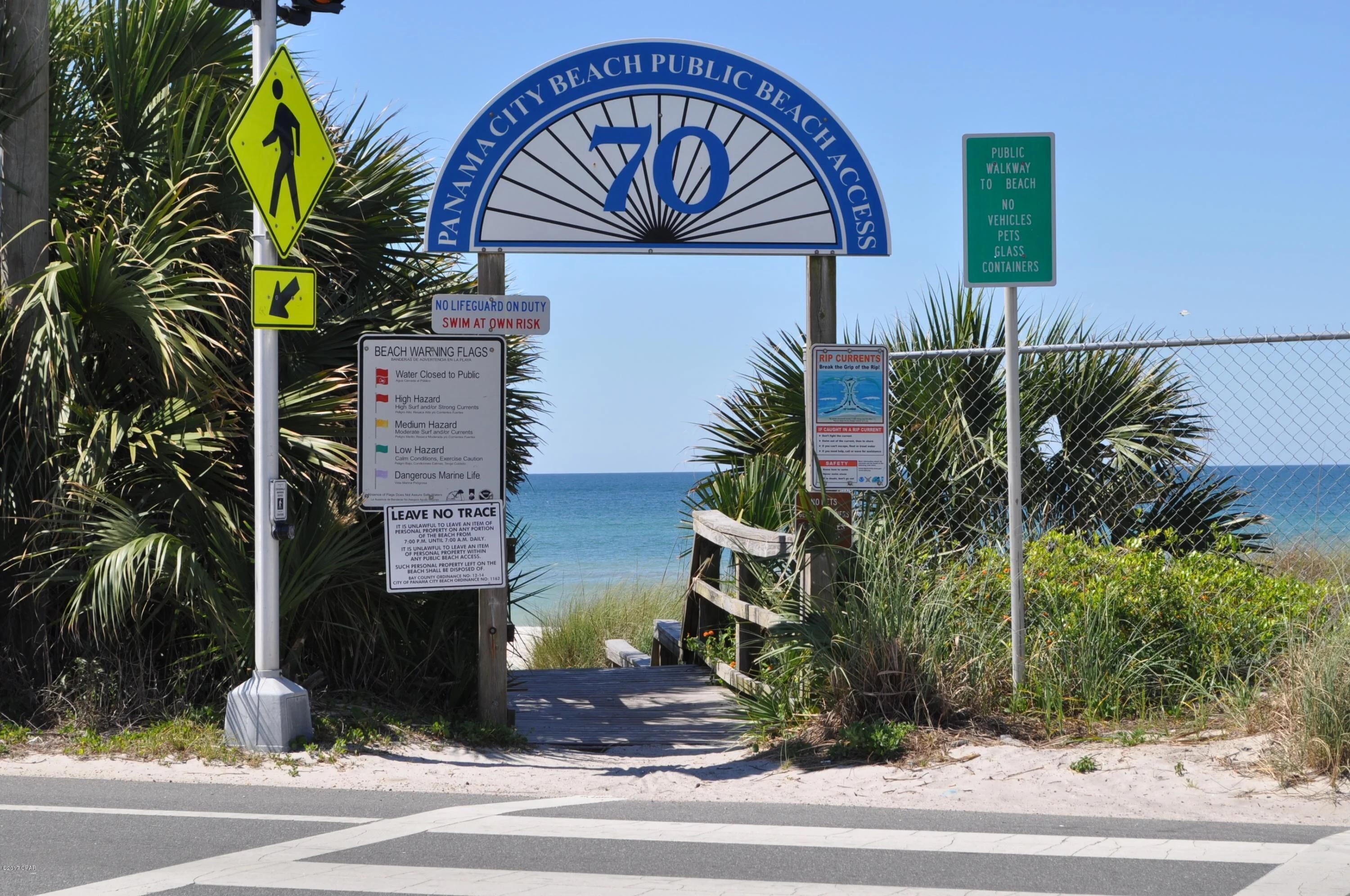 A beach entrance sign with a lifeguard on duty, beach warning flags, and a public walkway sign.