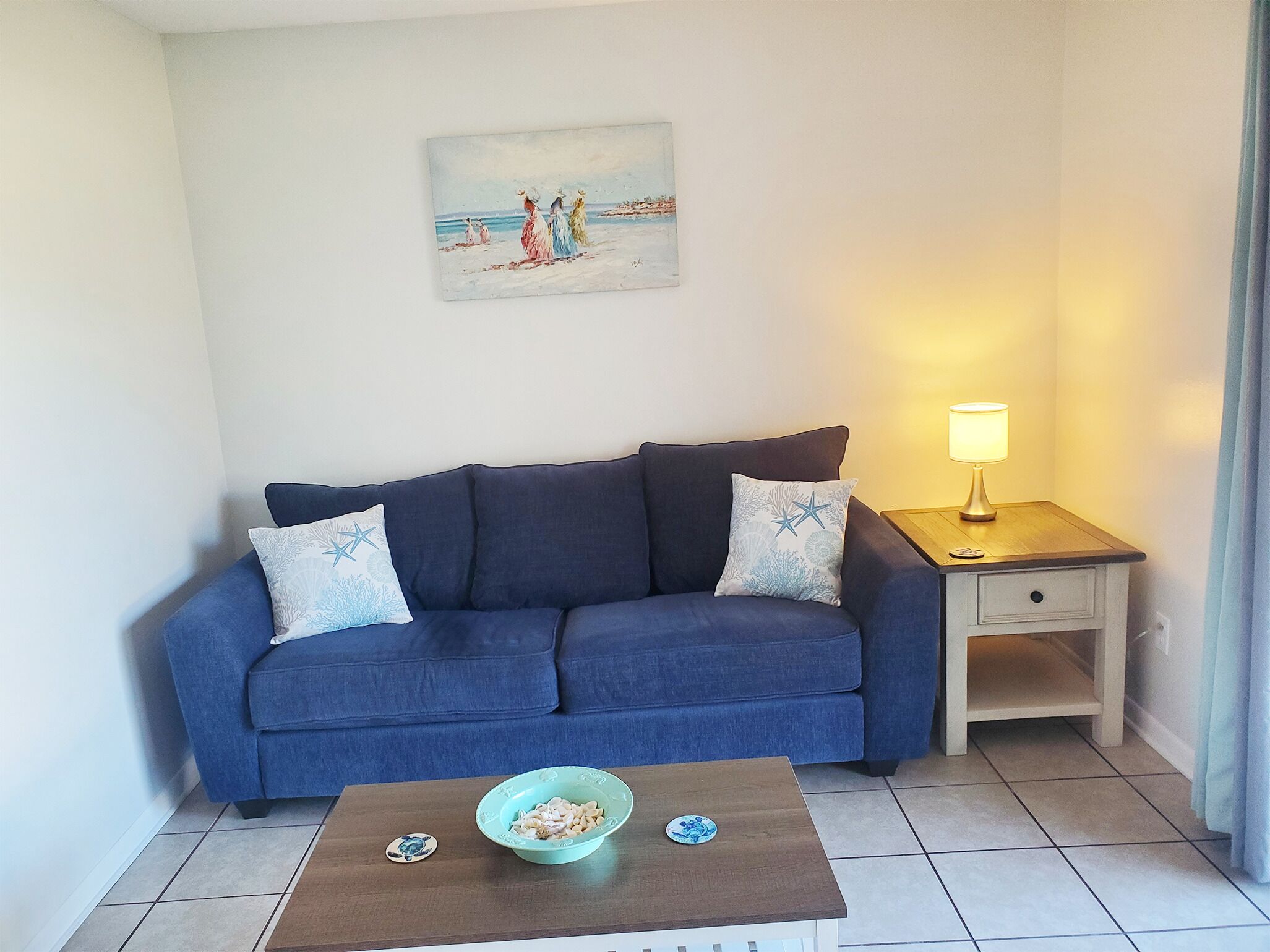 Ett vardagsrum med en blå soffa, två dekorativa kuddar, ett litet bord med en skål och tallrikar, en lampa och en tavla på väggen.