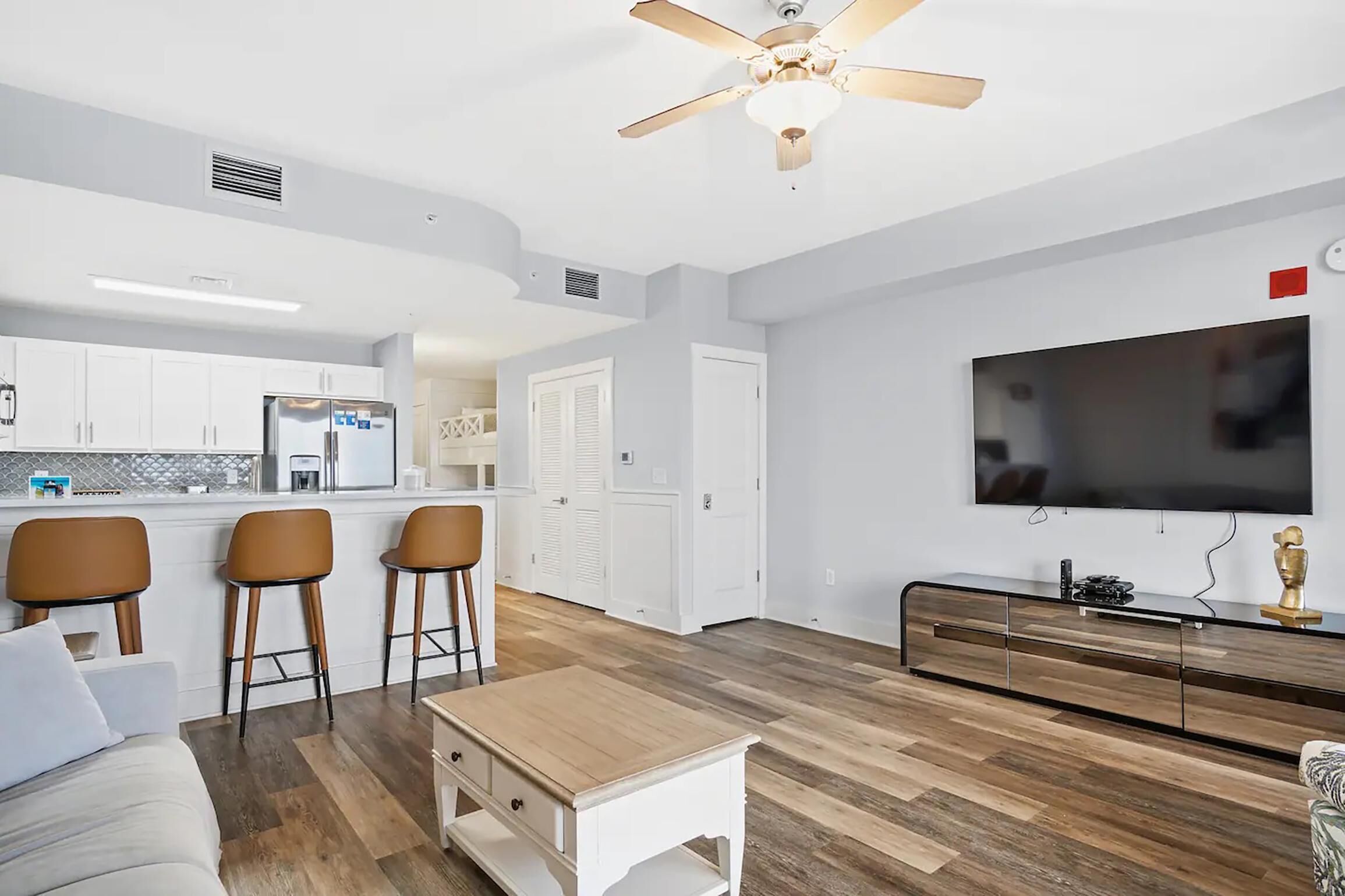 A modern living room with a sofa, coffee table, and a TV. There is a kitchen area with white cabinets and a refrigerator.