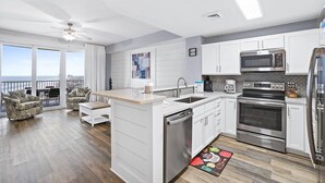 A modern kitchen with white cabinets, stainless steel appliances, and a large island. There is a dining area with a view of the ocean. - Calypso 3-2210 - Love Nest (Panama City Beach)