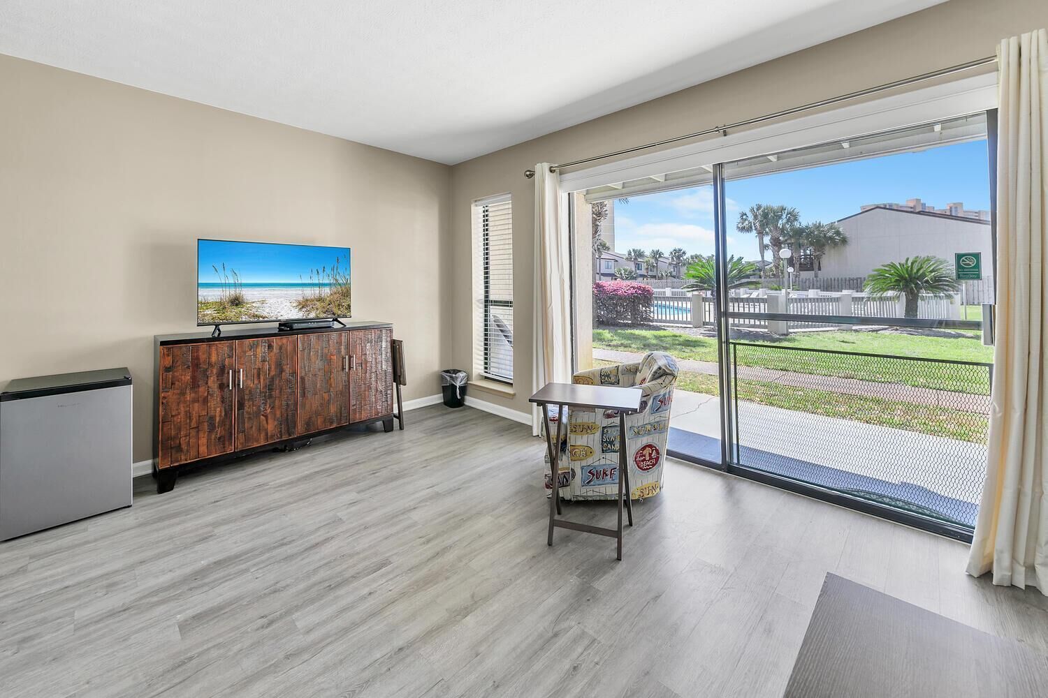 Una habitación con un armario de madera, una mesita y una silla. Se observa un ventanal con vistas a una playa y palmeras.