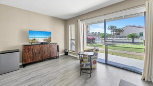 A room with a wooden cabinet, a small table, and a chair. There is a large window with a view of a beach and palm trees. - Endless Summer E2 (Panama City Beach)