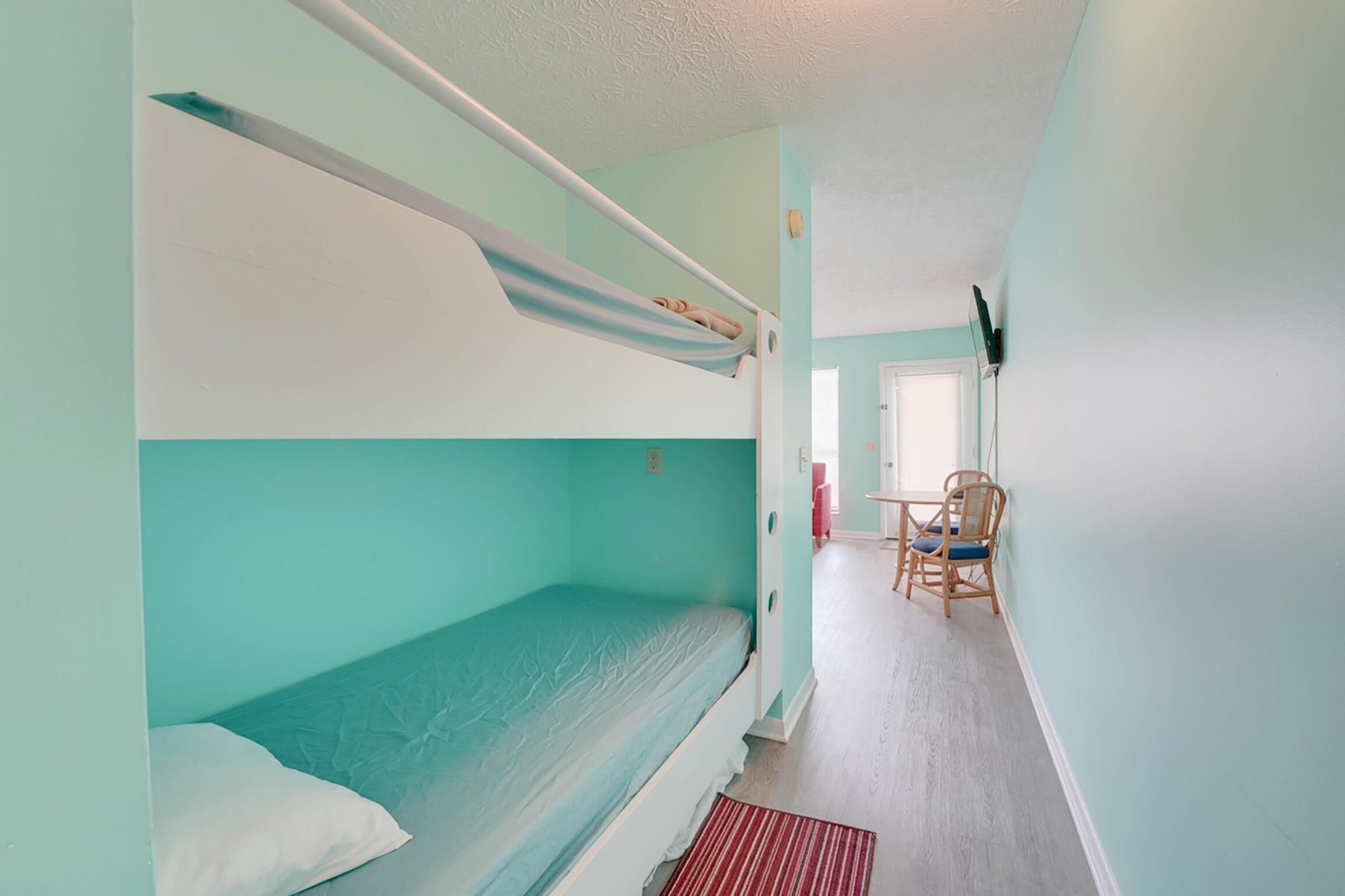 Een slaapkamer met een stapelbed, bureau en stoel, een rood tapijt en een deur naar een andere kamer.