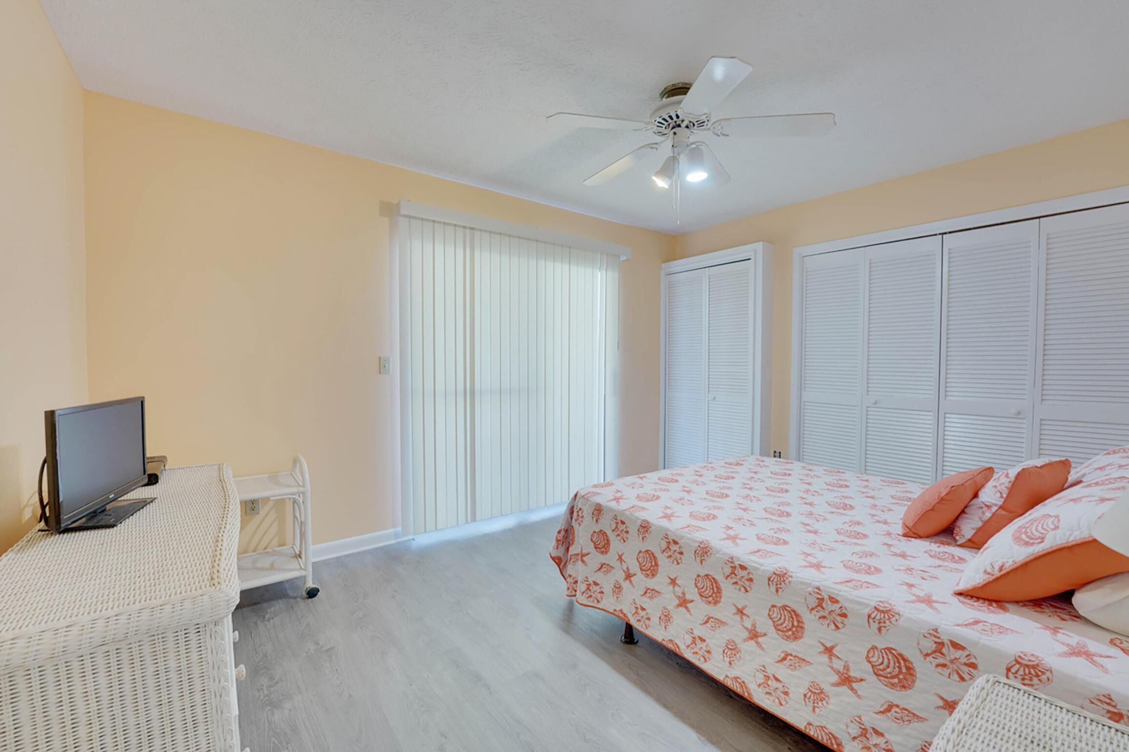 A bedroom with a bed, a television on a stand, a ceiling fan, and a window with blinds.