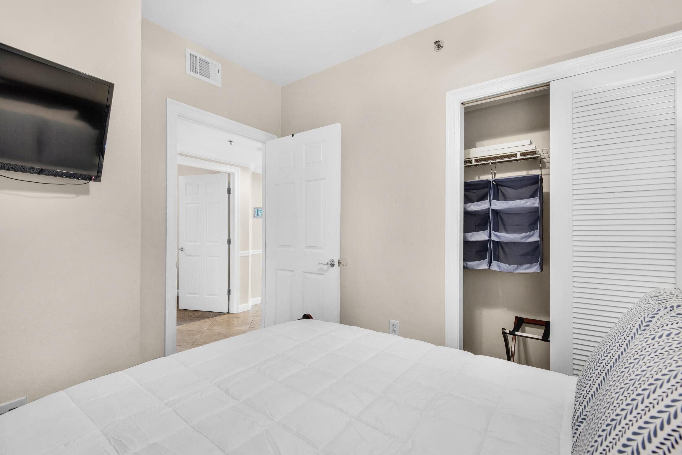 A bedroom with a bed, a closet with hanging clothes, a television on the wall, and a door leading to another room.