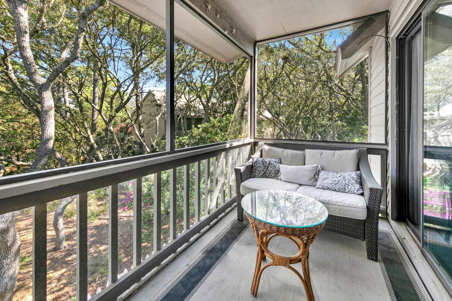 A balcony with a wicker sofa, a glass table, and a view of trees and houses.