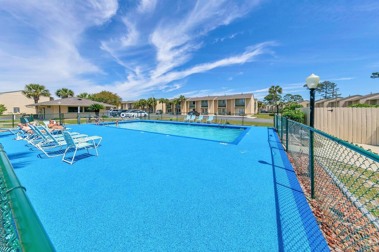 A swimming pool with lounge chairs, surrounded by a fence and a building in the background.