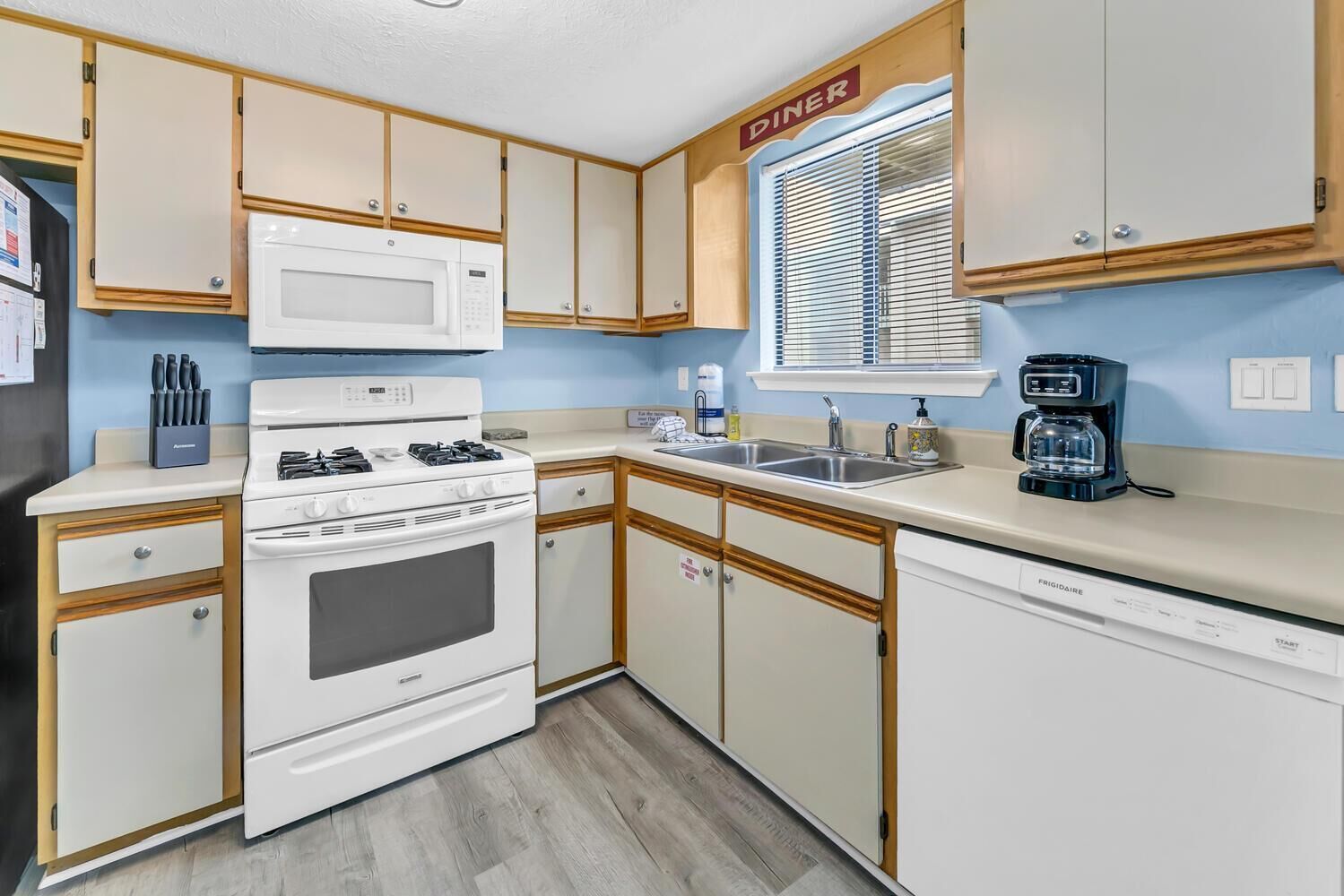 A compact kitchen with a white stove, microwave, and dishwasher, wooden cabinets, and a window with blinds.