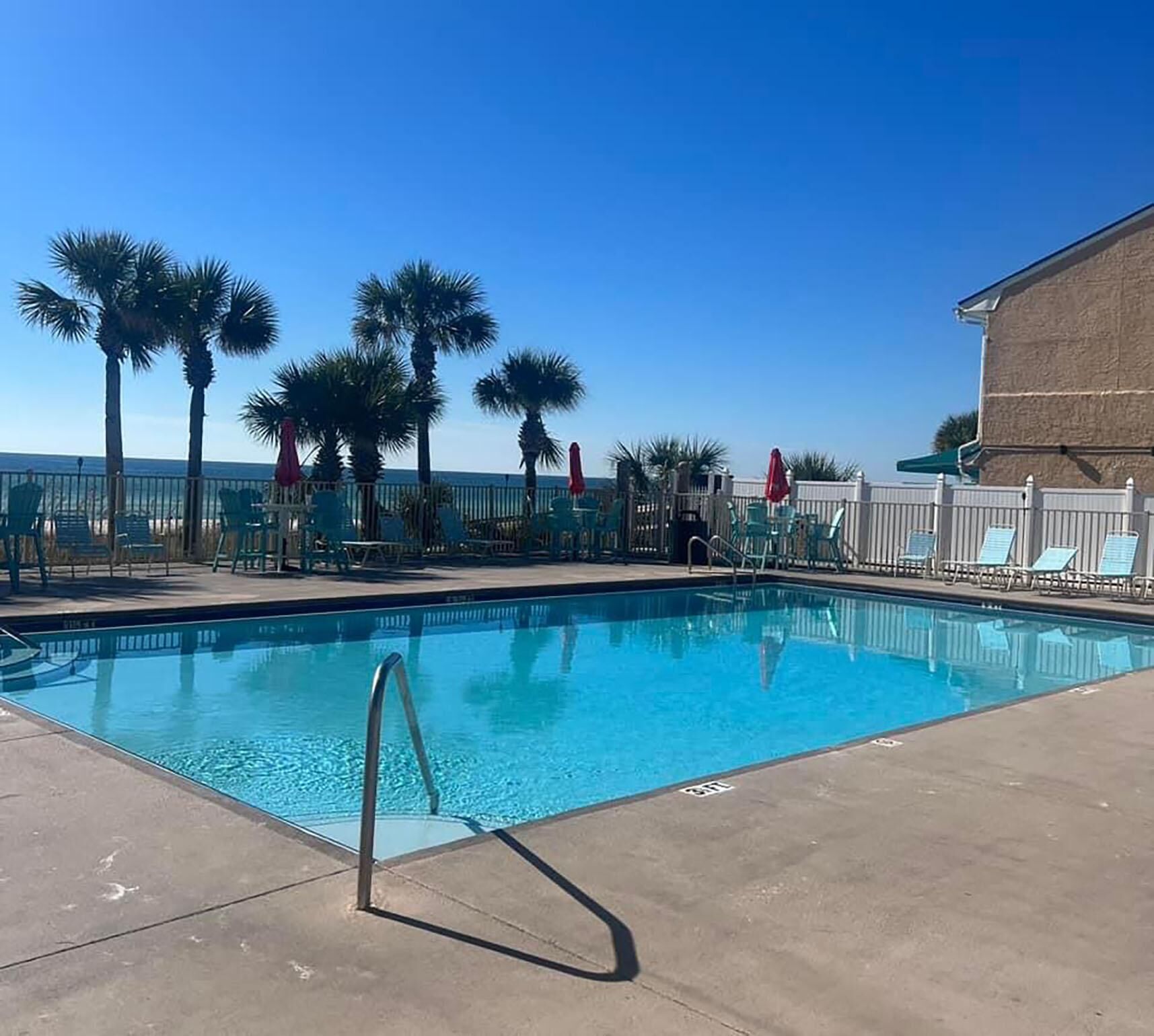 A swimming pool with a metal ladder, surrounded by palm trees and lounge chairs, with a building in the background.