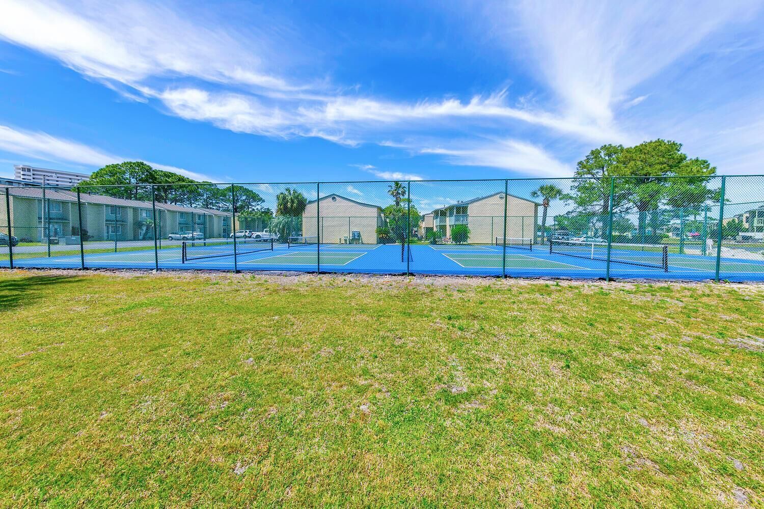 A tennis court with a chain-link fence, surrounded by grass and trees.