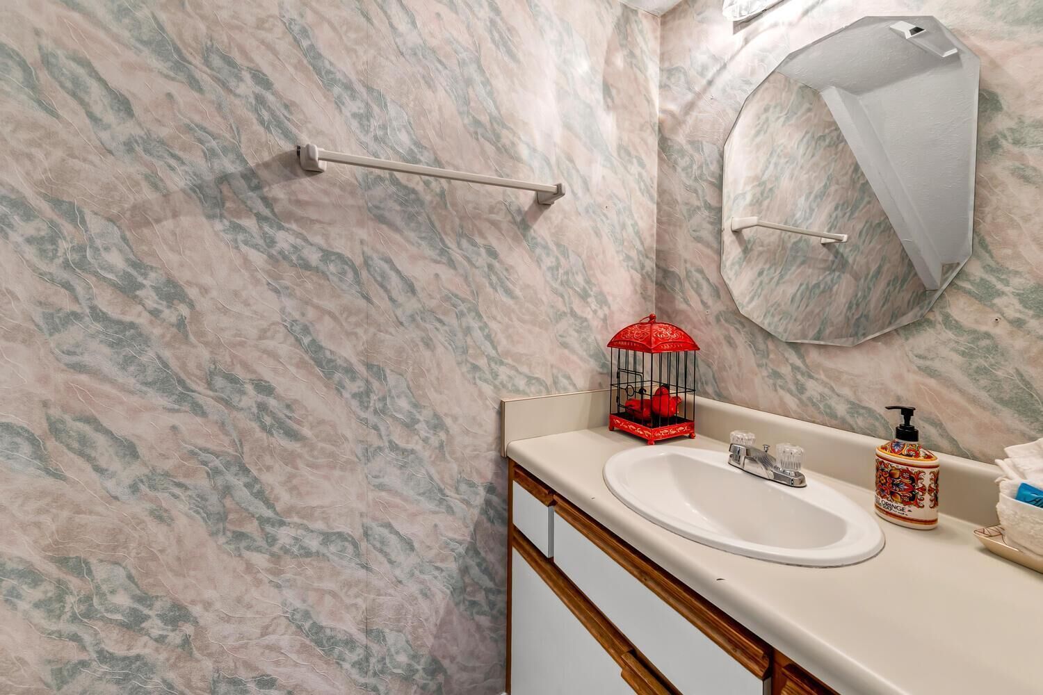 A bathroom with a marble wall, a sink, a mirror, a towel rack, a red birdcage, and a soap dispenser.
