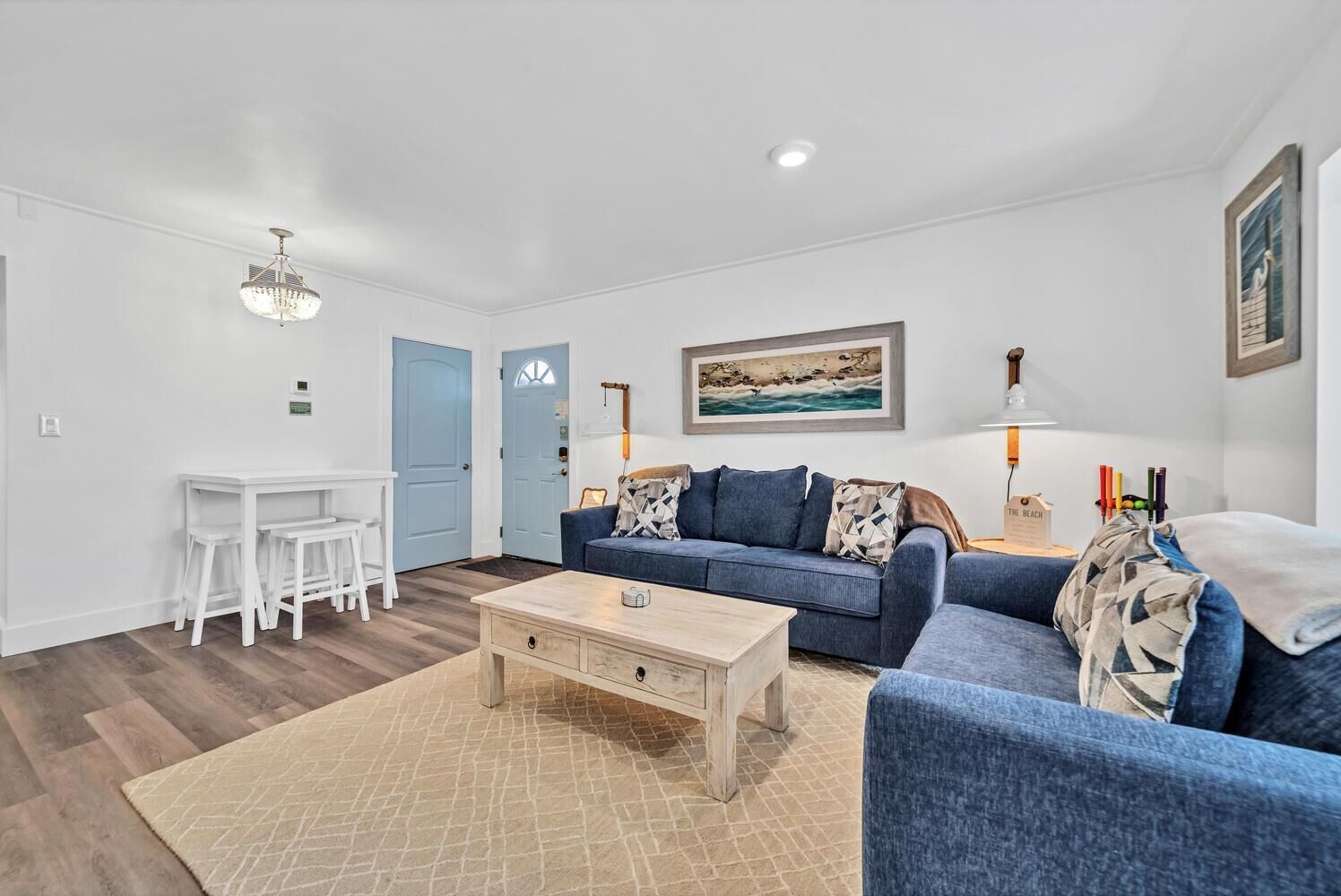 A living room with a blue sofa, a wooden coffee table, a white desk with stools, and a painting on the wall.