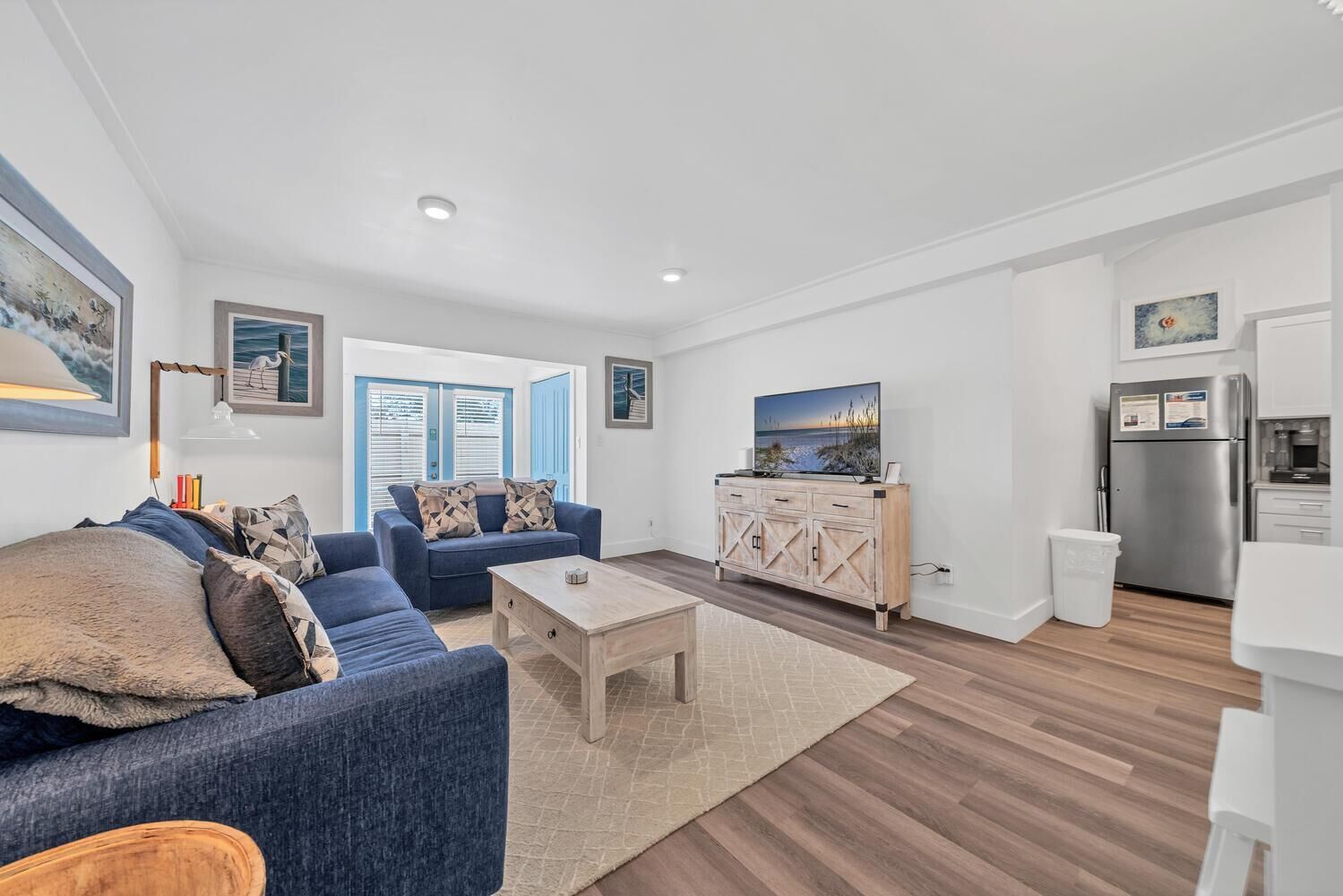 A modern living room with a blue sofa, a wooden coffee table, and a kitchen area with a refrigerator and microwave.