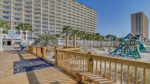 Un hotel sul lungomare con una passerella in legno che conduce a una terrazza piscina, un'area giochi per bambini e un grande edificio sullo sfondo.