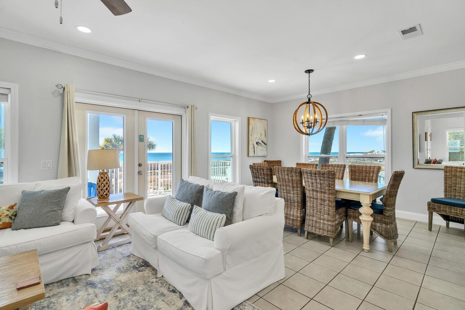 A spacious living area with a white sofa set, a dining table with wicker chairs, and large windows offering a view of the beach.