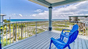 A wooden deck with a blue chair, overlooking a beach with a flag, and a building in the background. - 3 Generations Beach House (Santa Rosa Beach)