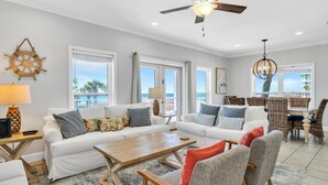 A spacious living room with a white sofa, a wooden coffee table, and wicker chairs. There is a ceiling fan, large windows with a view, and a dining area with wicker furniture. - 3 Generations Beach House (Santa Rosa Beach)