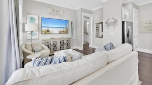 A spacious living room with a large sofa, a television, and a cabinet. There is a sign on the wall and a door leading to another room. - Town of Prominence 67C - Pour Beach Life (Santa Rosa Beach)