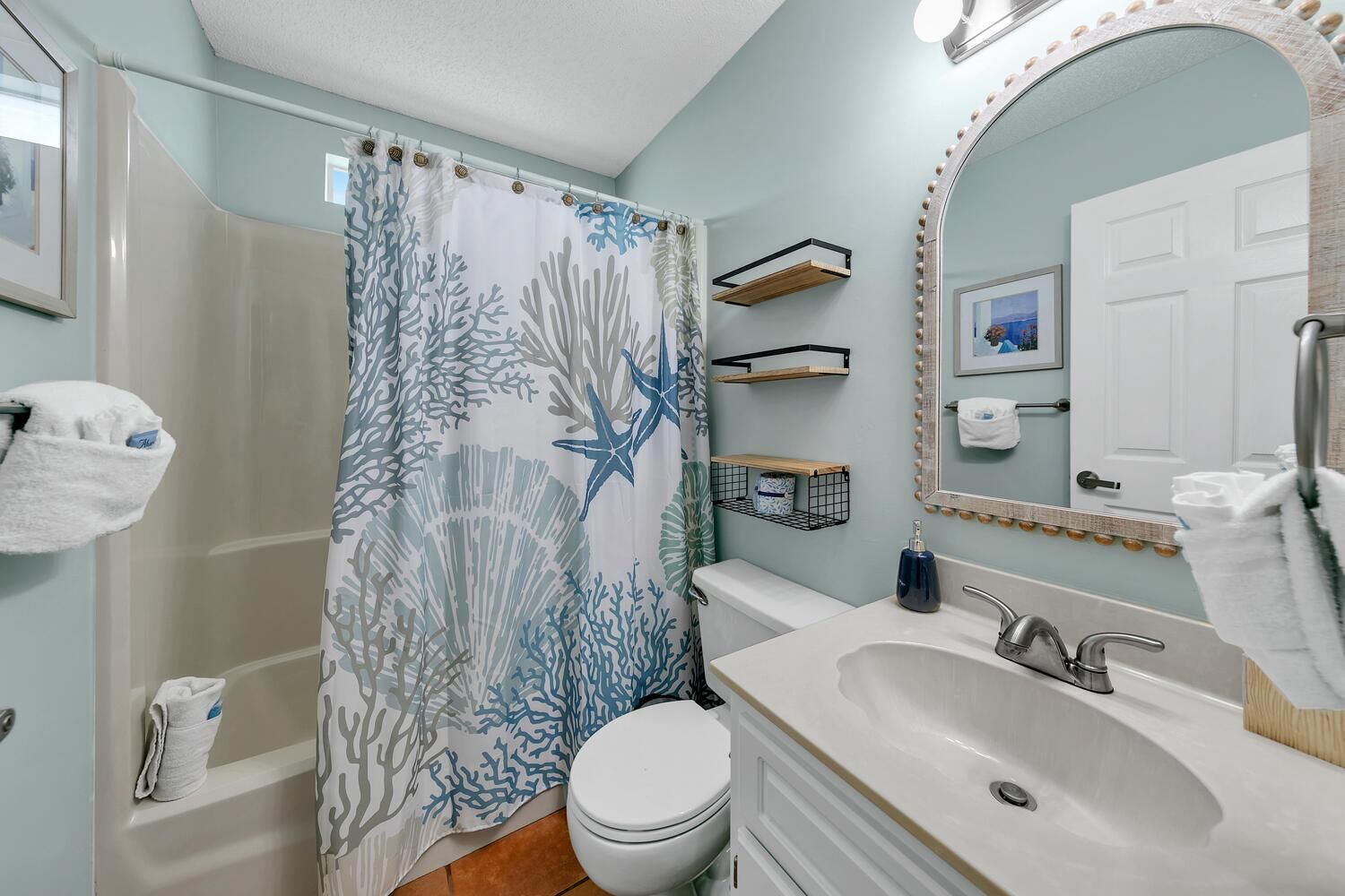 A bathroom with a shower curtain featuring a coral and starfish pattern, a white toilet, a sink with a mirror, and a towel rack.