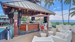 Ein überdachter Außenbereich mit Bar, einem Pool, weißen Sitzgelegenheiten mit Polstern und Meerblick.