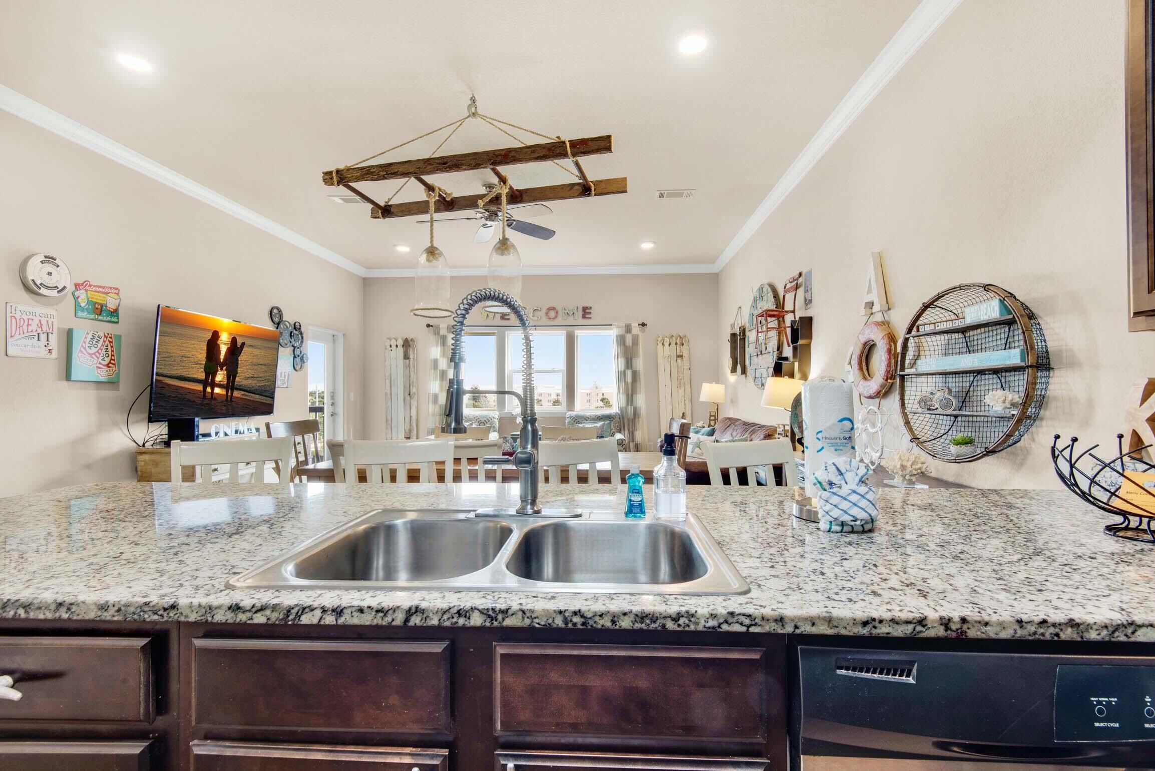 A kitchen with granite countertops, stainless steel sinks, and a built-in dishwasher. There is a ceiling fan, a hanging light fixture, and a welcome sign on the wall.