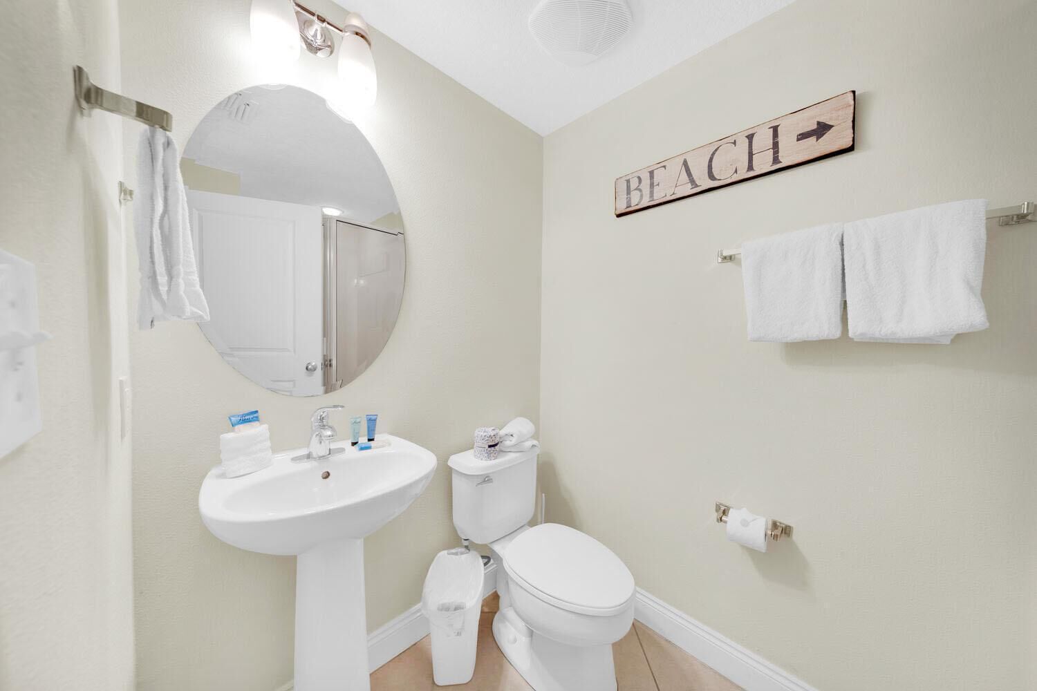 Ein Badezimmer mit Waschbecken, Toilette und Spiegel. An der Wand befindet sich ein Schild mit der Aufschrift 'BEACH' und einem Pfeil, der nach rechts zeigt.