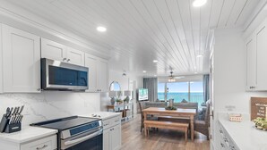 A modern kitchen with white cabinetry, a built-in microwave, and an oven. There is a dining area with a wooden table and chairs, and a view of the ocean through large windows. - Beach House 502d (Miramar Beach)