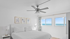 A bedroom with a bed, a ceiling fan, a nightstand, a lamp, and a view of the ocean through large windows. - Beach House 502d (Miramar Beach)