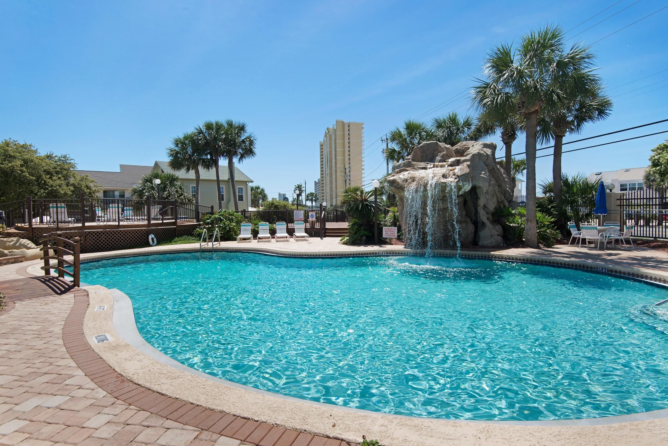 A swimming pool with a waterfall feature, surrounded by palm trees and lounge chairs.