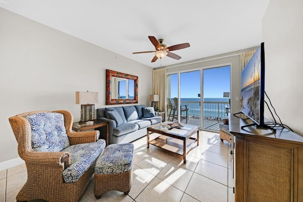 A spacious living room with a sofa, two armchairs, a coffee table, a TV, and a balcony with a view of the ocean.