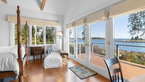 A bedroom with a wooden bed, a nightstand, a dresser, and a view of a lake through large windows. - Chimerique on the Water in Maine (Saint George)